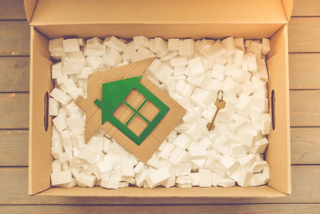 A brown cardboard moving box is open, showing white packing peanuts, a gold key, and a green house shape.